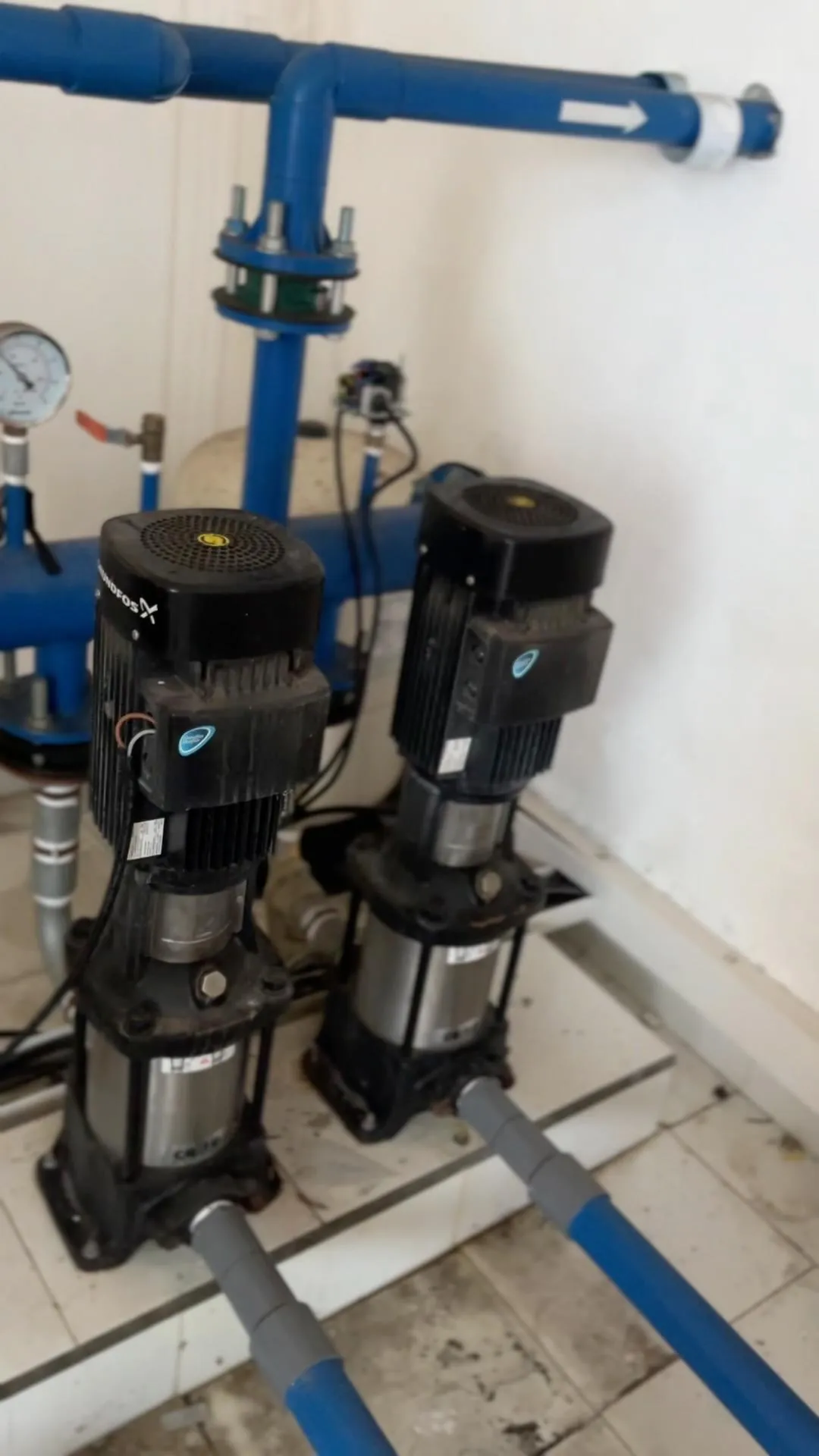 Dua unit pompa air vertikal dengan instalasi pipa biru, manometer, dan tangki di ruang pompa